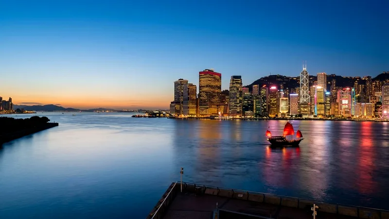 Hong Kong Island skyline at blue hour across calm Victoria Harbour with a red-sailed junk.